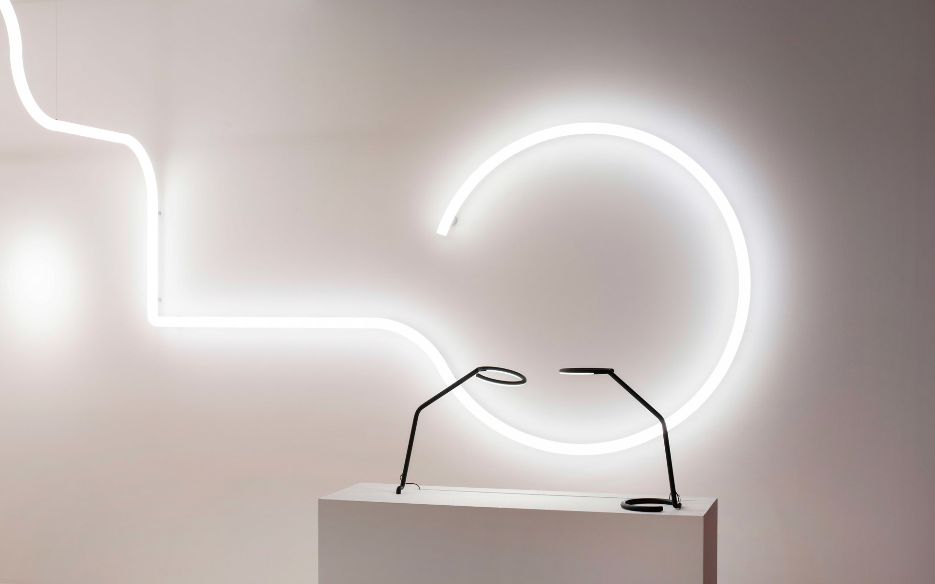 Artemide flexible LED light tubes forming curves on a wall, with a sleek black desk lamp on a white pedestal below