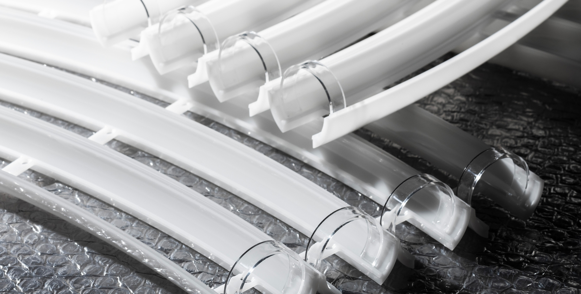 Profili estrusi curvi bianchi e trasparenti per apparecchi illuminotecnici, disposti su una superficie riflettente con pluriball.