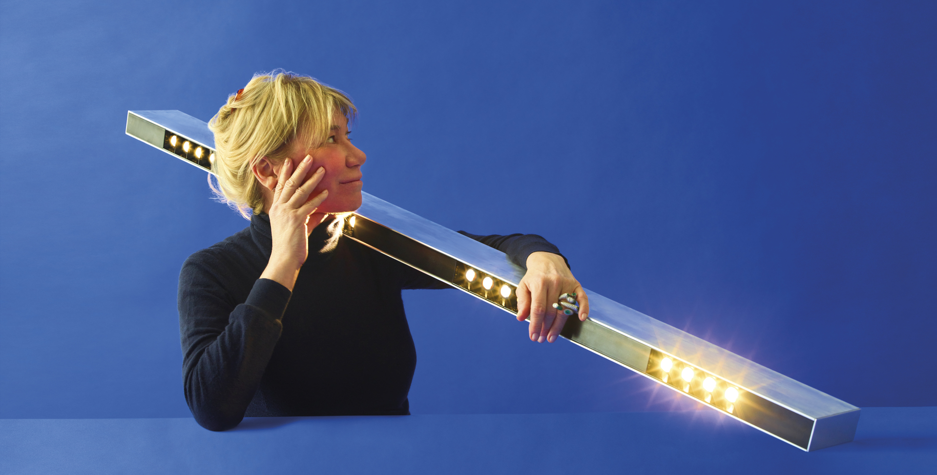 Donna che regge una lampada a barra LED lineare Artemide con faretti bianco caldo su sfondo blu.