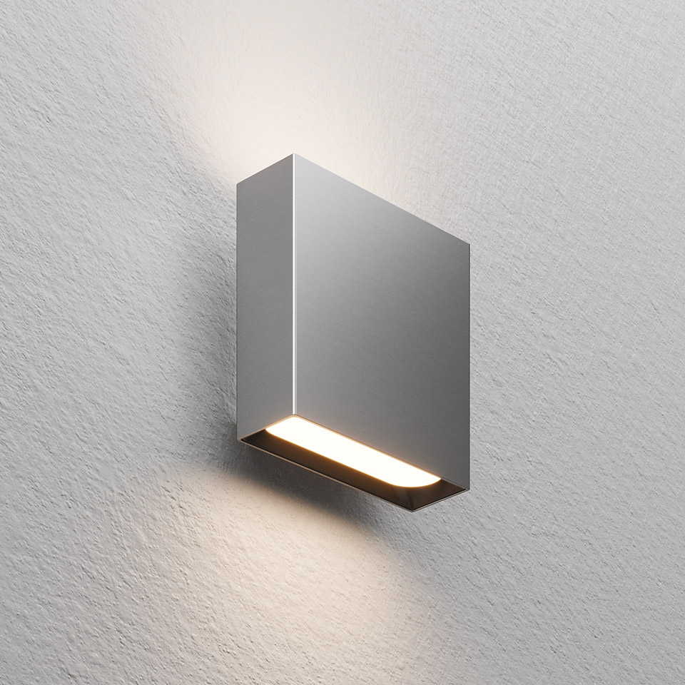 Applique Agathe di Artemide: corpo rettangolare grigio compatto con striscia LED inferiore che emette luce calda verso il basso su parete testurizzata