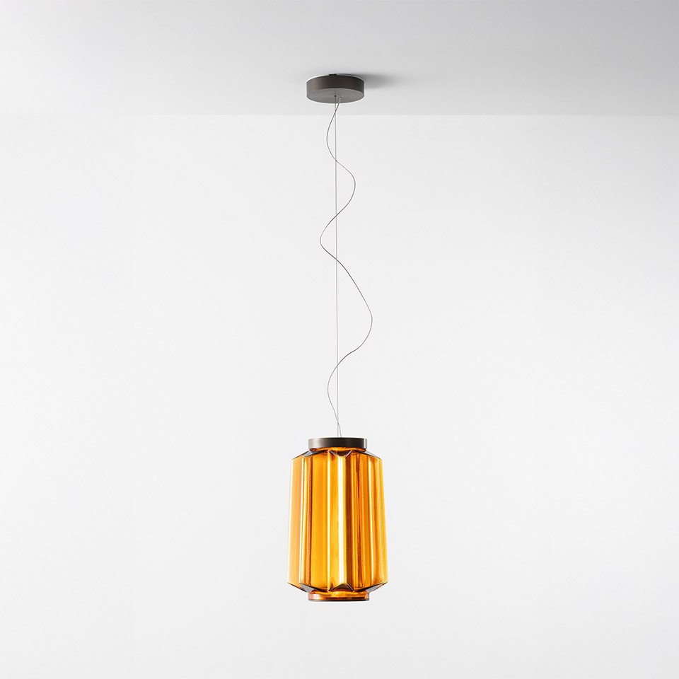 Lampada a sospensione Artemide Auralia con diffusore in vetro ambra a pannelli costolati verticali, sospesa da un sottile cavo a un rosone grigio a soffitto