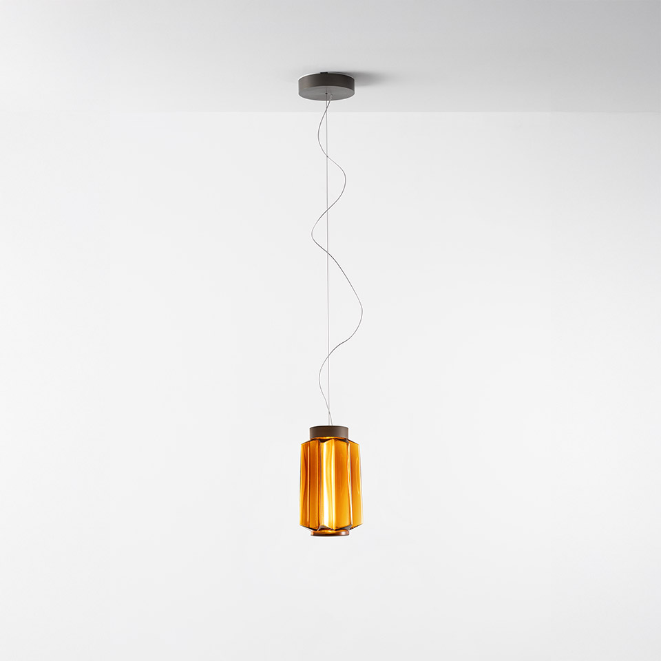 Lampada a sospensione Artemide Auralia con diffusore in vetro costolato cilindrico ambra e sommità in metallo scuro, sospesa da un cavo sottile
