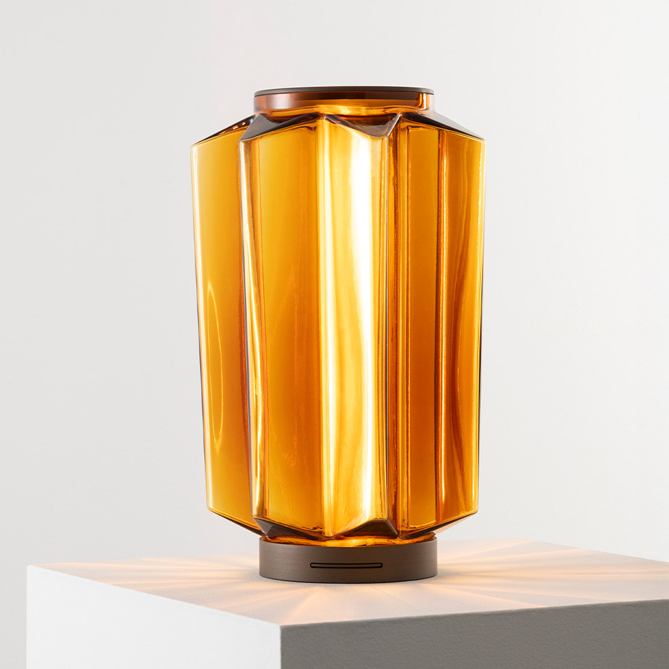 Lampada da tavolo Auralia di Artemide: diffusore in vetro ambrato sfaccettato con luce calda, base in metallo bronzo con batteria ricaricabile