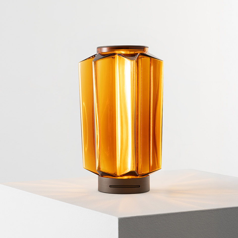 Lampada da tavolo Auralia di Artemide: diffusore in vetro ambrato sfaccettato con base bronzo, emette luce dorata calda su superficie bianca