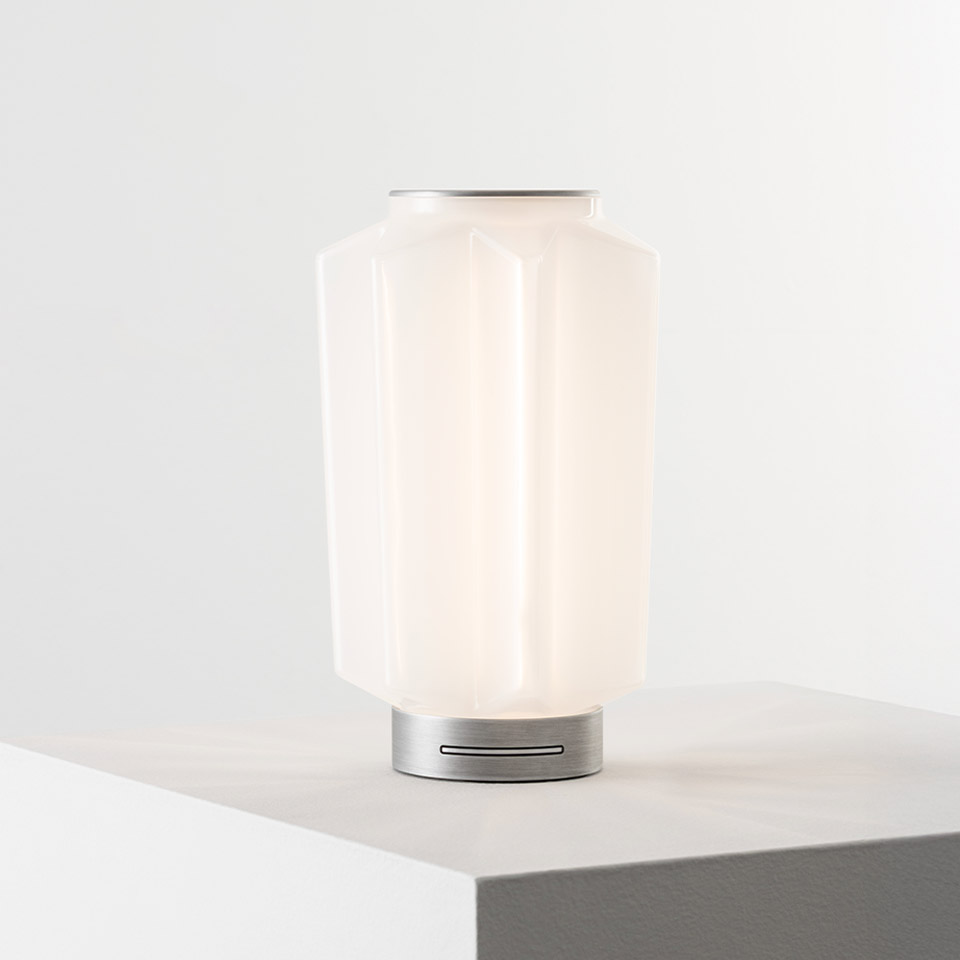 Lampada da tavolo Artemide Auralia con diffusore in vetro bianco opalino e base in alluminio spazzolato, illuminata delicatamente su una superficie