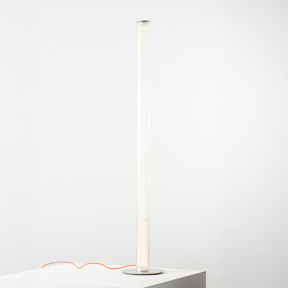 Artemide Criosfera floor lamp: tall slender cylindrical mesh diffuser emitting soft white light, on a round base with orange cord