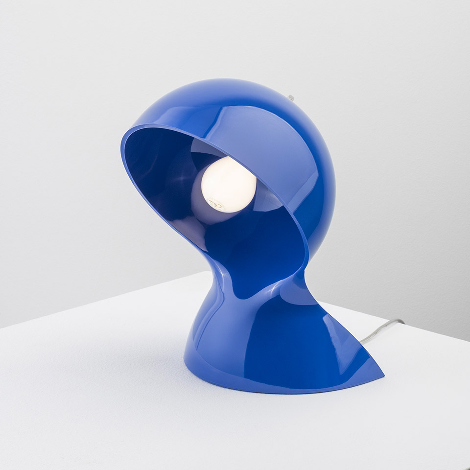 Lampada da tavolo Artemide Dalù in blu lucido, con paralume scultoreo a forma di casco e lampadina a vista, su superficie bianca