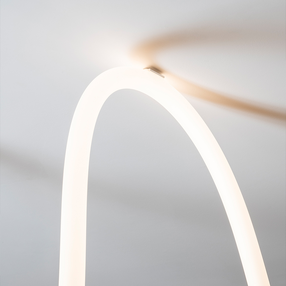 Primo piano della lampada Artemide La Linea: un arco tubolare LED bianco luminoso che emette luce diffusa calda su sfondo grigio chiaro