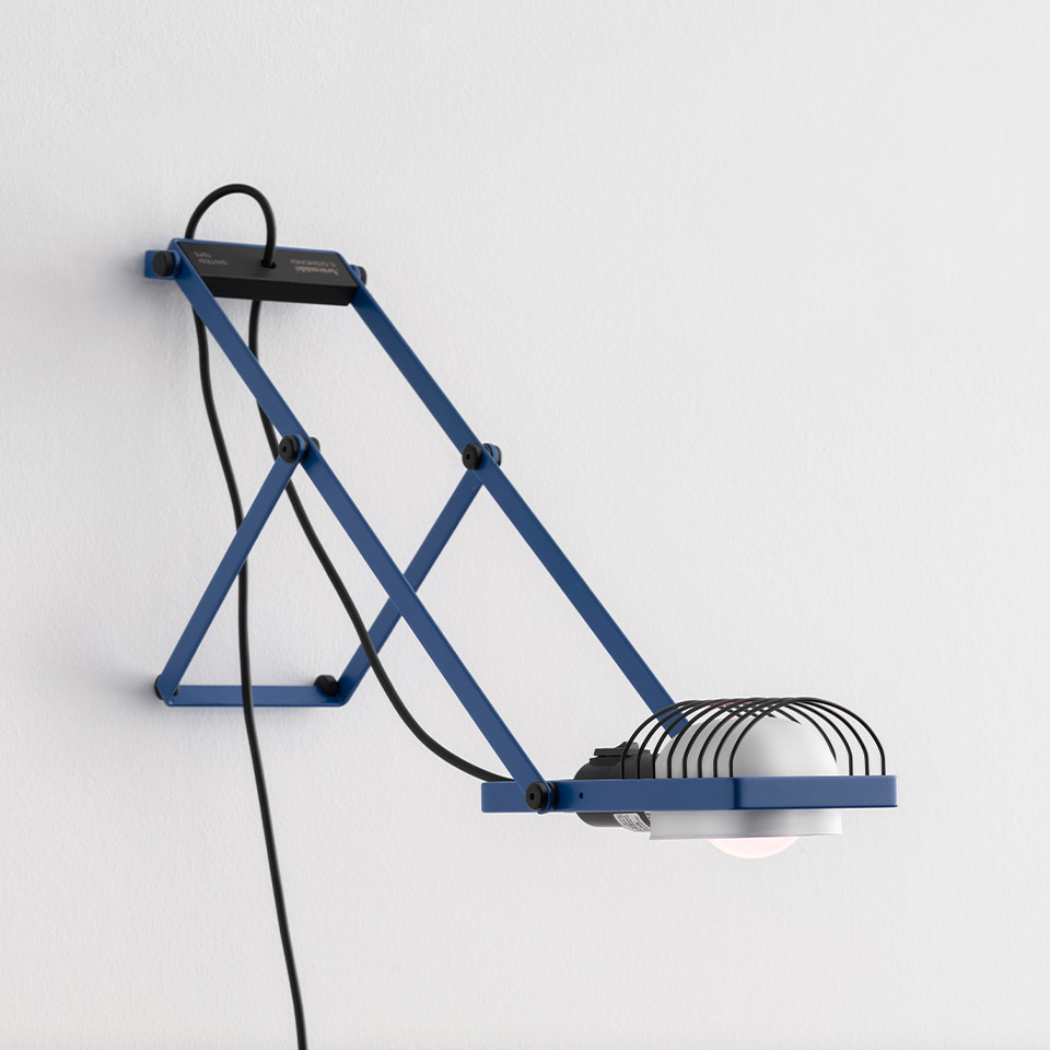 Lampada da parete Sintesi blu con meccanismo a pantografo, paralume a gabbia metallica e cavo nero a vista, montata su parete bianca
