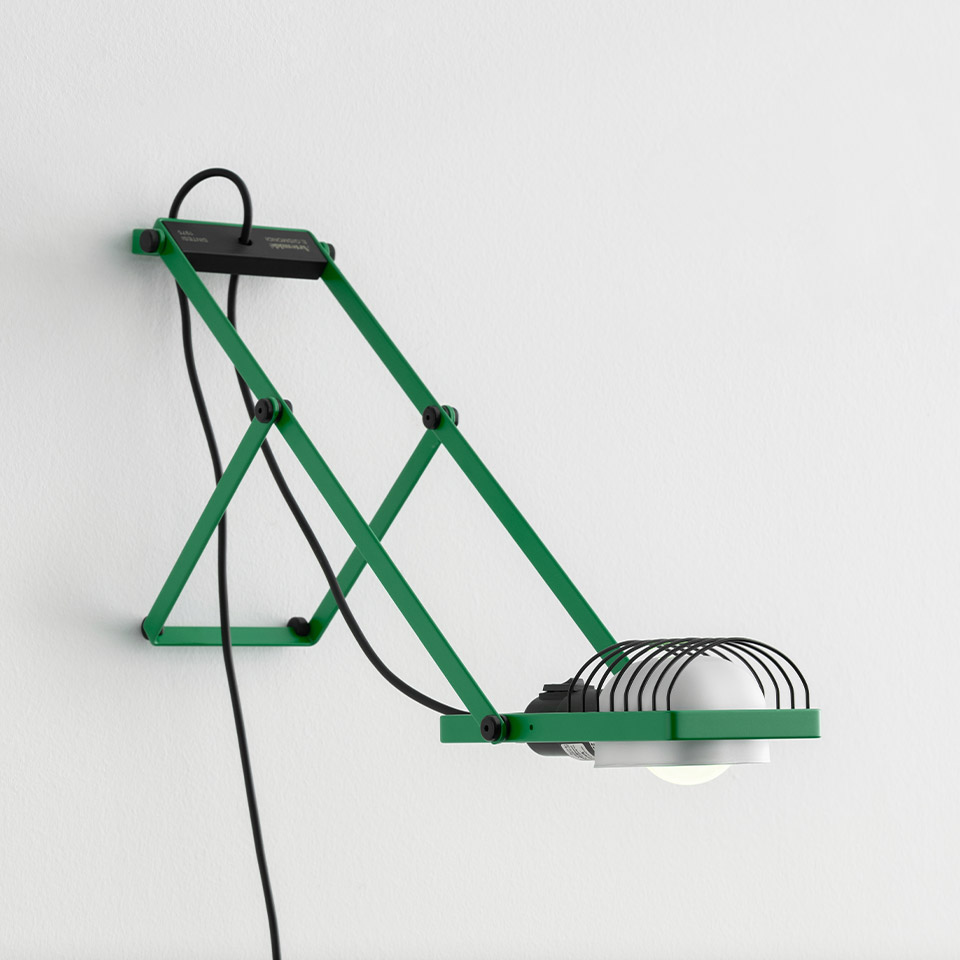 Lampada da parete Sintesi di Artemide: struttura a pantografo verde con paralume a griglia metallica, bulbo a vista e cavo nero su parete bianca