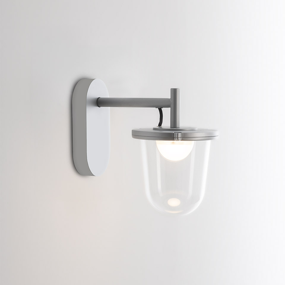 Applique da esterno Artemide Tolomeo Outdoor: braccio in metallo grigio su piastra bianca, paralume a campana in vetro trasparente con bulbo opale visibile