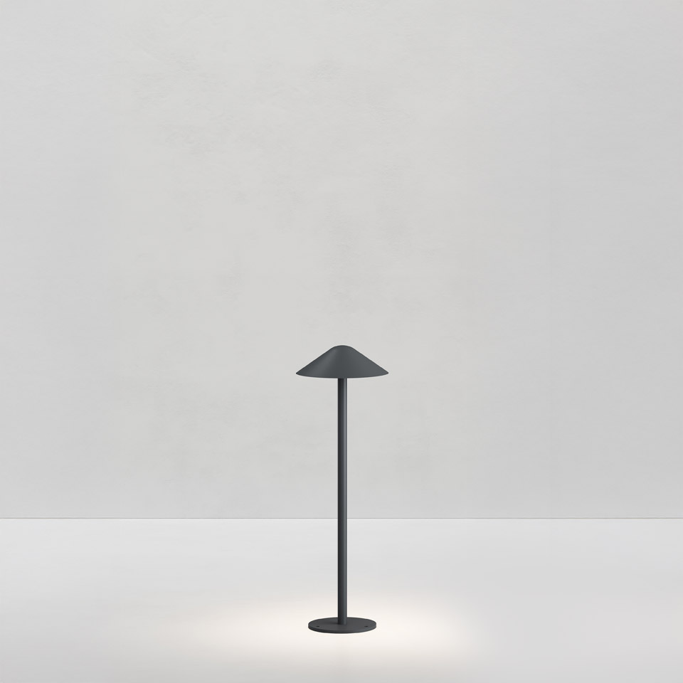 Lampada da tavolo Artemide Vea Bollard in grigio scuro, stelo sottile con paralume conico, emette luce soffusa verso il basso su sfondo bianco