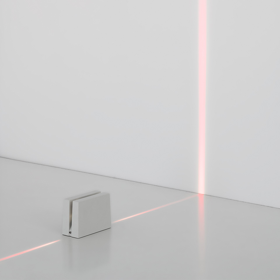 Lampada da terra Antarktikós di Artemide: cubo bianco compatto che emette una precisa linea di luce rossa sul pavimento e sulla parete
