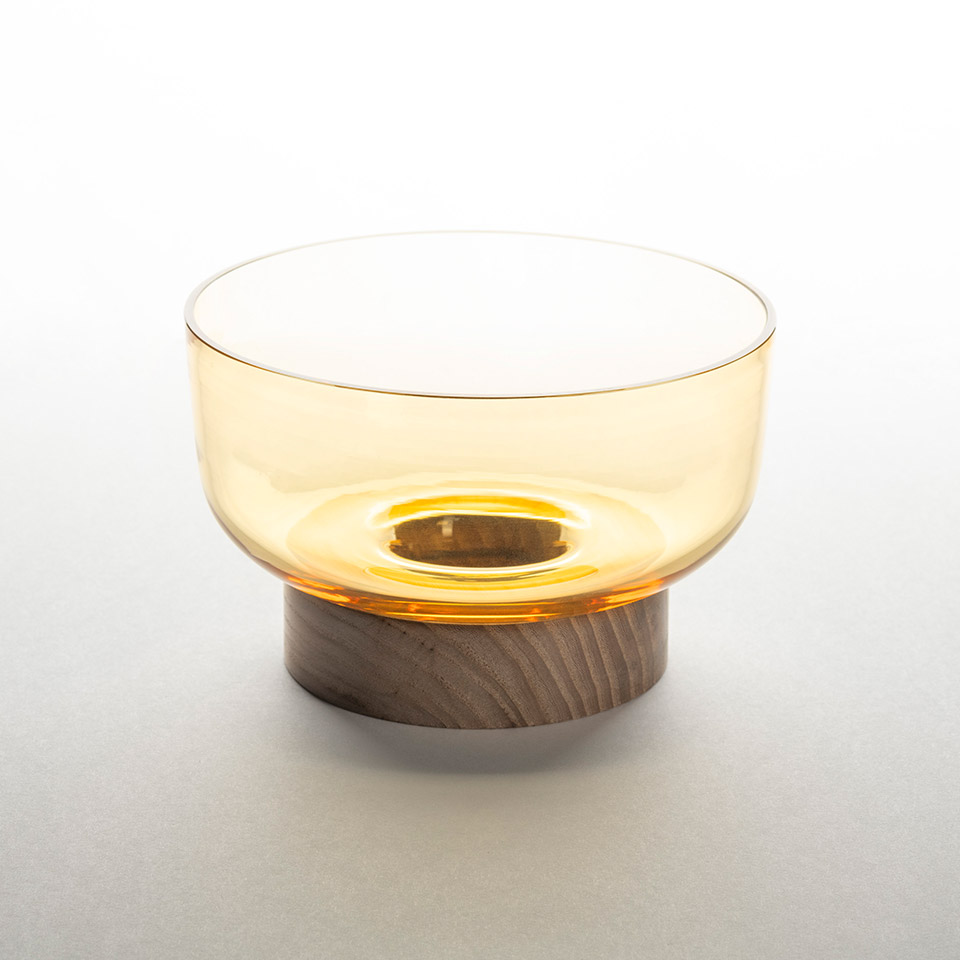 Artemide Bontà table lamp: amber transparent glass bowl diffuser resting on a cylindrical natural wood base, emitting warm light