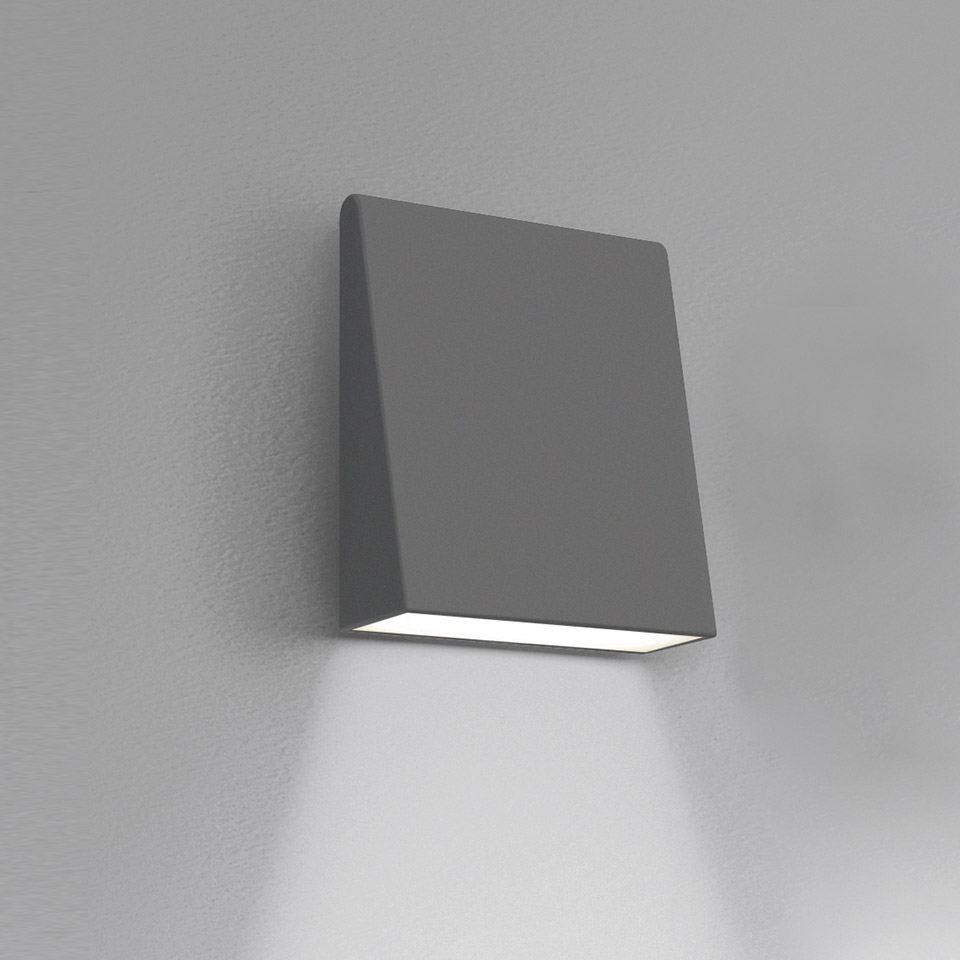 Applique Artemide Cuneo, corpo quadrato compatto grigio, emette fascio di luce bianca verso il basso, montata su parete grigia