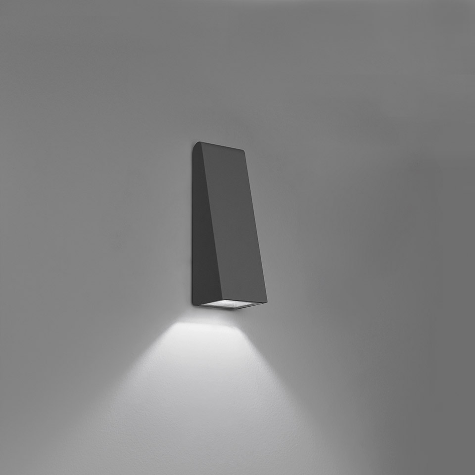 Lampada da parete Artemide Cuneo, corpo trapezoidale antracite scuro, con fascio LED orientato verso il basso su sfondo neutro