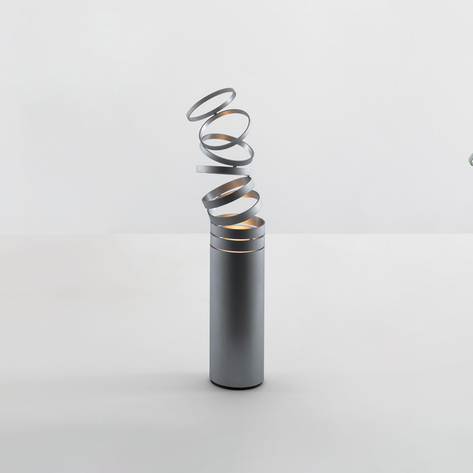 Artemide Decomposé Light Tavolo: base cilindrica grigia con diffusore a nastro metallico a spirale verso l'alto che emette luce calda