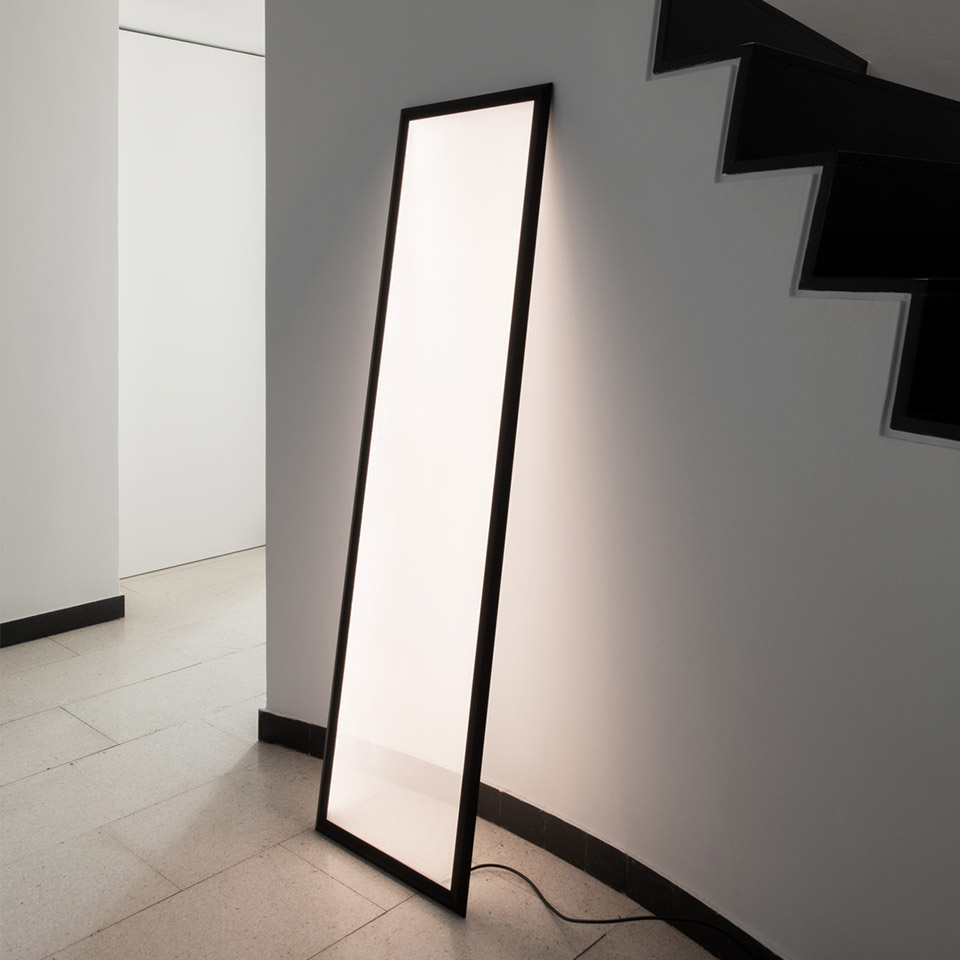 Lampada da terra Artemide Discovery Terra: alto pannello luminoso rettangolare con cornice nera, appoggiato alla parete vicino a una scala, emette luce bianca diffusa