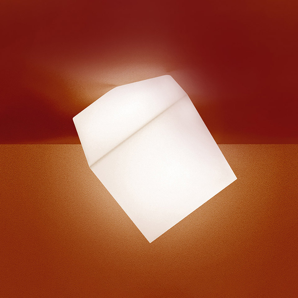 Artemide Edge lamp: tilted cube-shaped white opal diffuser emitting warm light against a red wall and orange surface