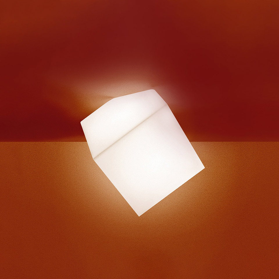 Artemide Edge wall/ceiling lamp: tilted opal white glass cube diffuser emitting warm light against a deep red wall and surface