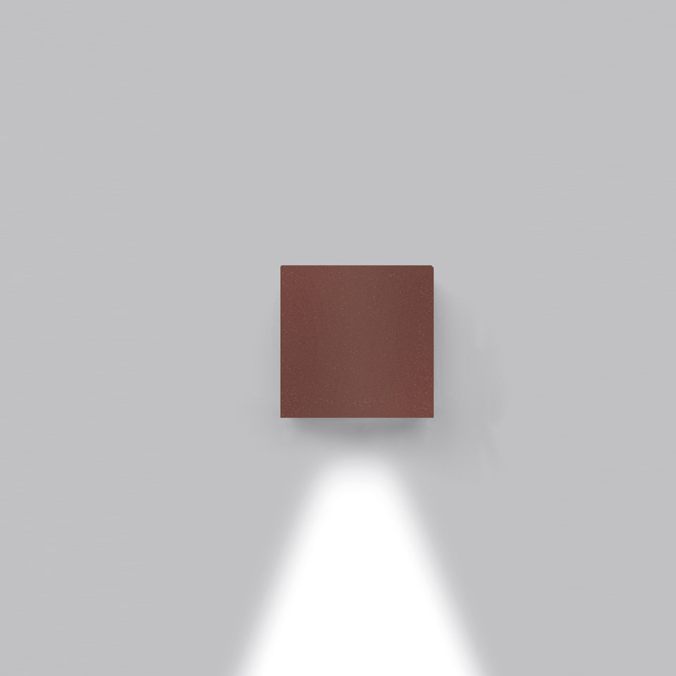 Applique Effetto di Artemide: corpo quadrato compatto color ruggine che emette un fascio di luce bianca diretto verso il basso su parete grigia