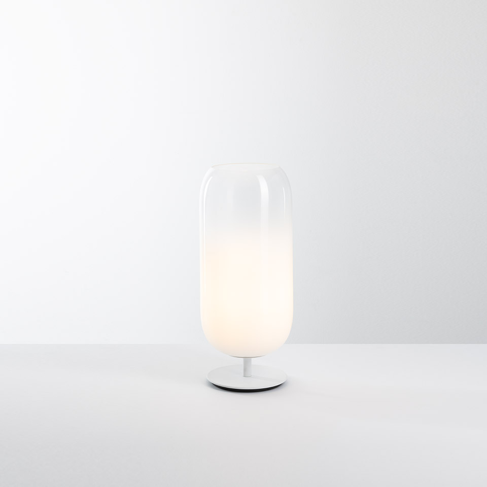 Artemide Gople table lamp: tall capsule-shaped opal white glass diffuser on a slim white metal base, emitting soft warm light - 2