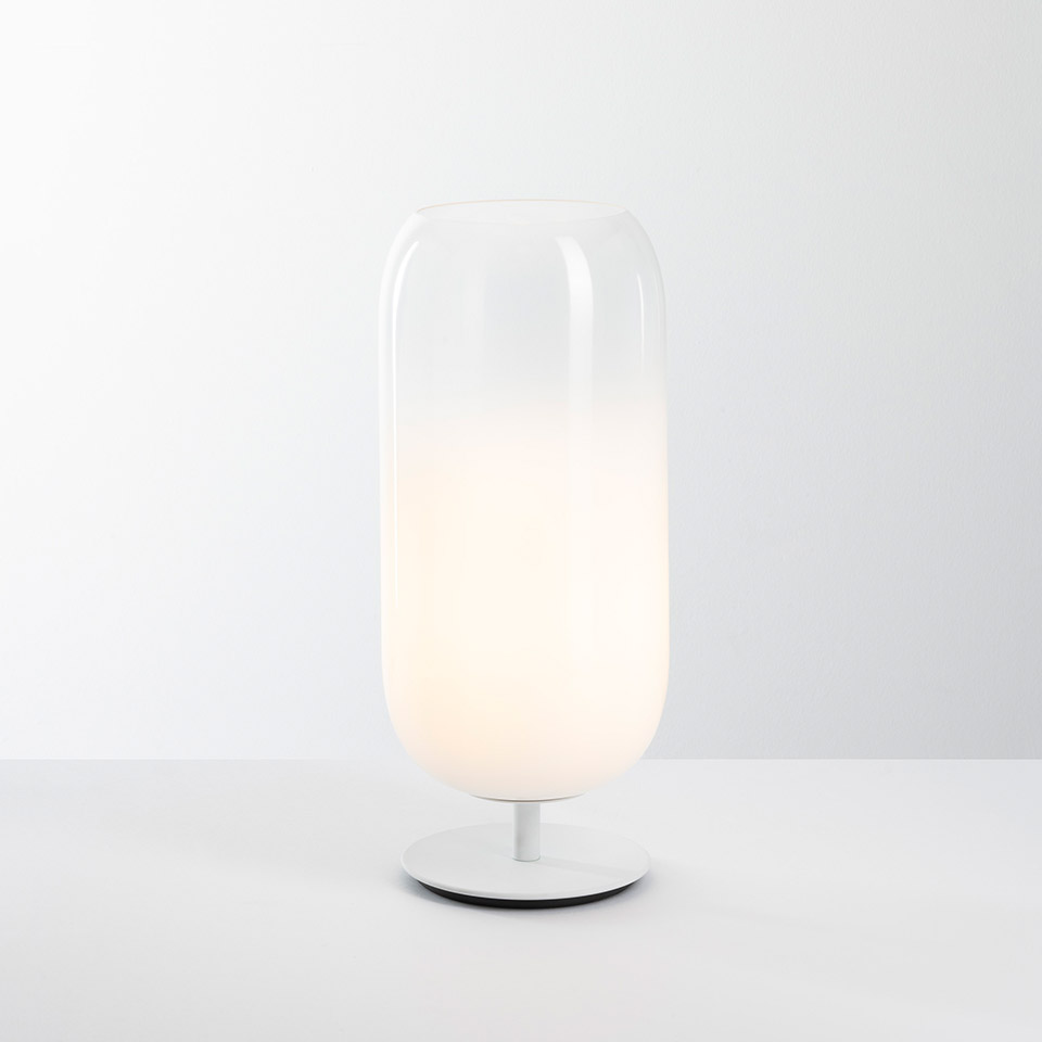 Artemide Gople table lamp: tall capsule-shaped opal white glass diffuser on a slim white metal base, emitting soft warm light - 2