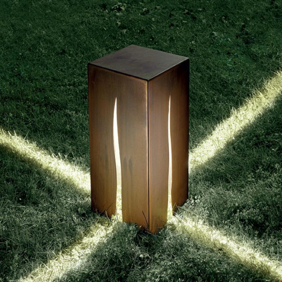 Lampada da esterno Artemide Granito in acciaio corten con fessure a fiamma che proiettano fasci di luce sull'erba