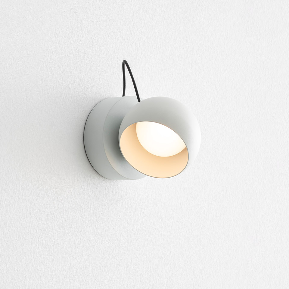 Lampada da parete Artemide Ixa: applique cilindrica bianca compatta con paralume rotondo orientabile, luce LED calda, cavo nero su parete bianca