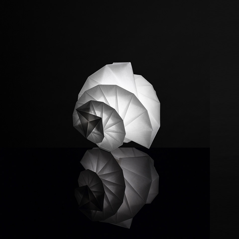 Artemide In-Ei Mendori di Issey Miyake: lampada da tavolo a spirale luminosa ispirata all'origami, forma di conchiglia bianca, riflessa su superficie nera