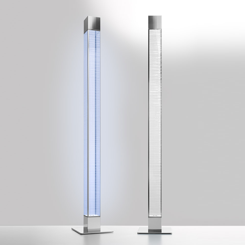Due lampade da terra Artemide Mimesi: sinistra accesa con luce blu-bianca, destra spenta, entrambe con colonna in cristallo e base quadrata cromata