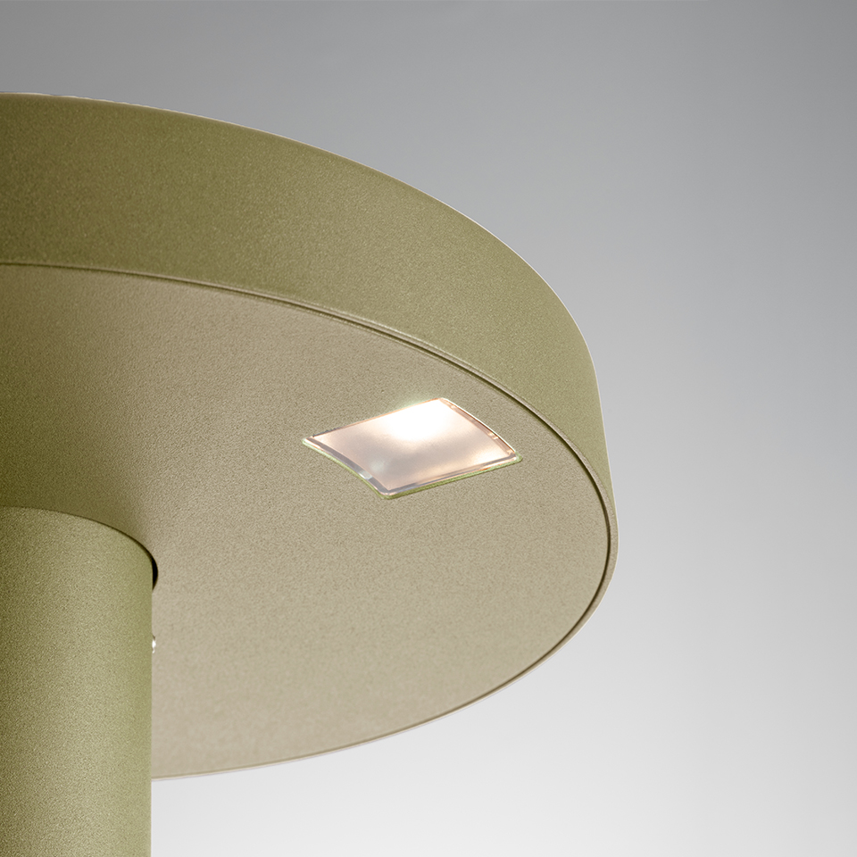 Primo piano della lampada Artemide Needoo: paralume circolare metallico oro-verde con piccolo pannello LED quadrato opaco sulla superficie superiore