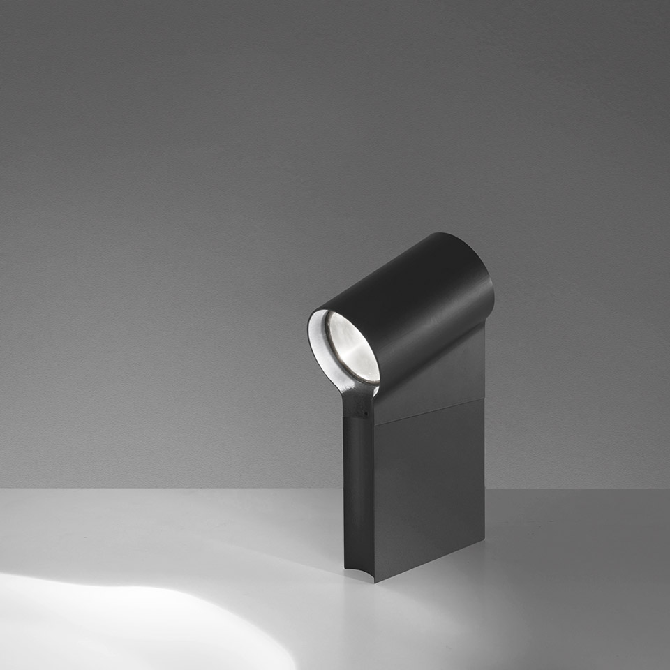 Lampada da tavolo Artemide Oblique nera, testa cilindrica inclinata in avanti su base piatta, proietta un fascio di luce concentrato