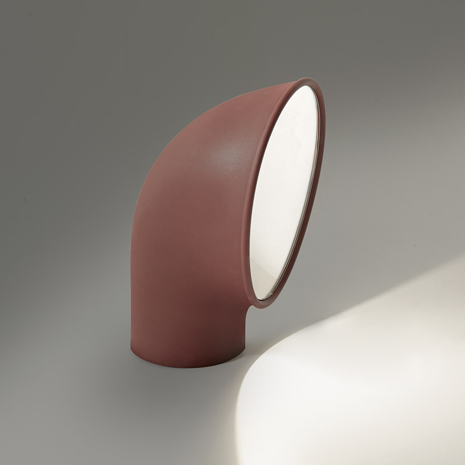 Artemide Piroscafo table lamp in terracotta red, curved tubular body angled upward, emitting a focused beam of light downward