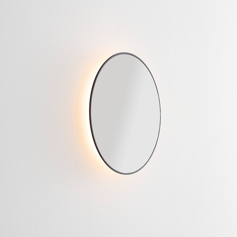 Specchio rotondo Artemide Se|eS con sottile cornice scura e retroilluminazione LED calda che risplende dolcemente su una parete bianca