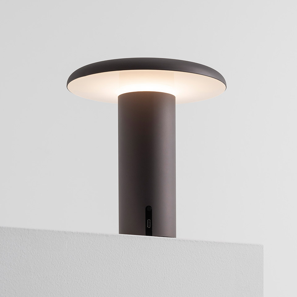 Artemide Takku portable LED table lamp in anodized dark brown, cylindrical stem with flat disc diffuser emitting warm white light