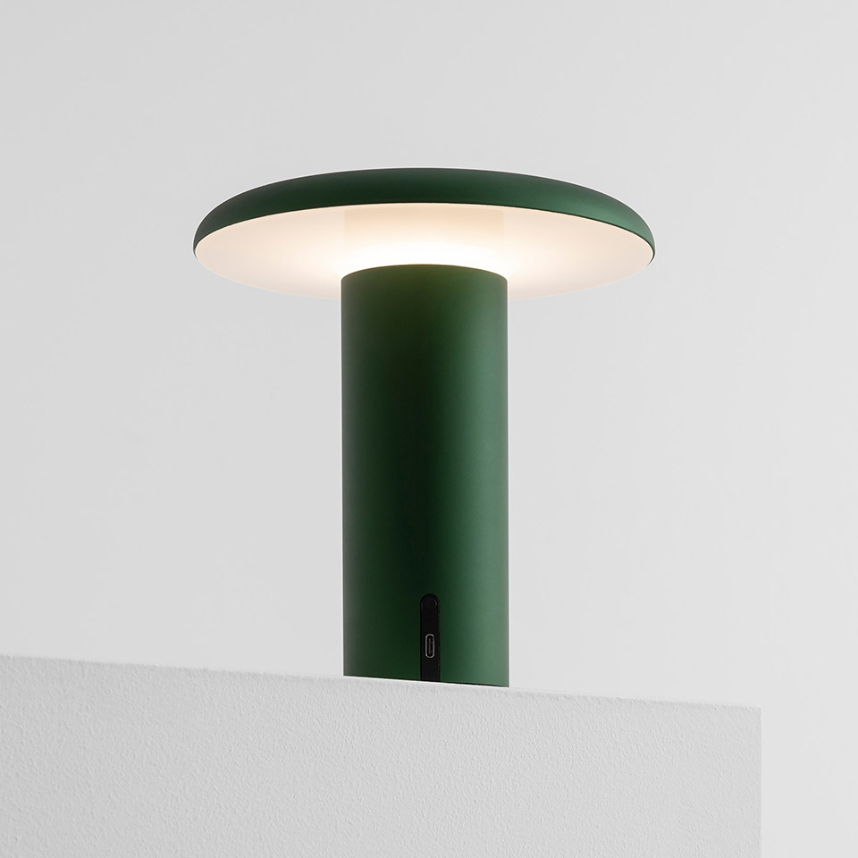 Artemide Takku portable LED table lamp in forest green, featuring a cylindrical base with flat mushroom-shaped diffuser emitting warm light