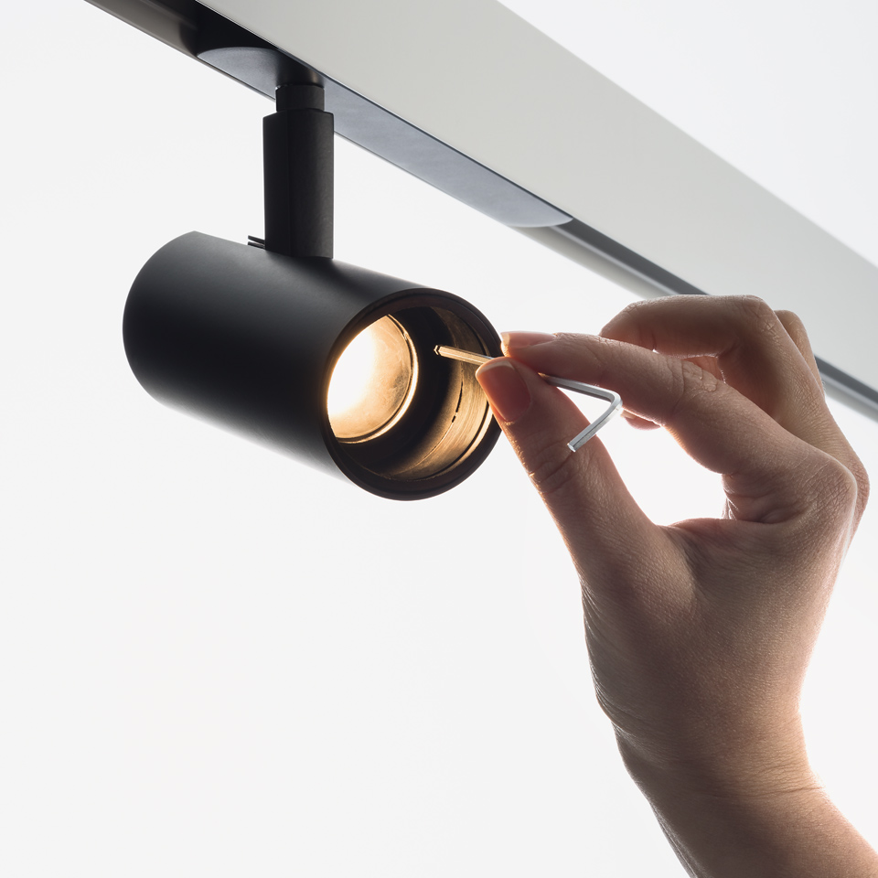 Mano che regola la lente di un faretto cilindrico nero Artemide Vector Track 230V montato su binario a soffitto. - 2