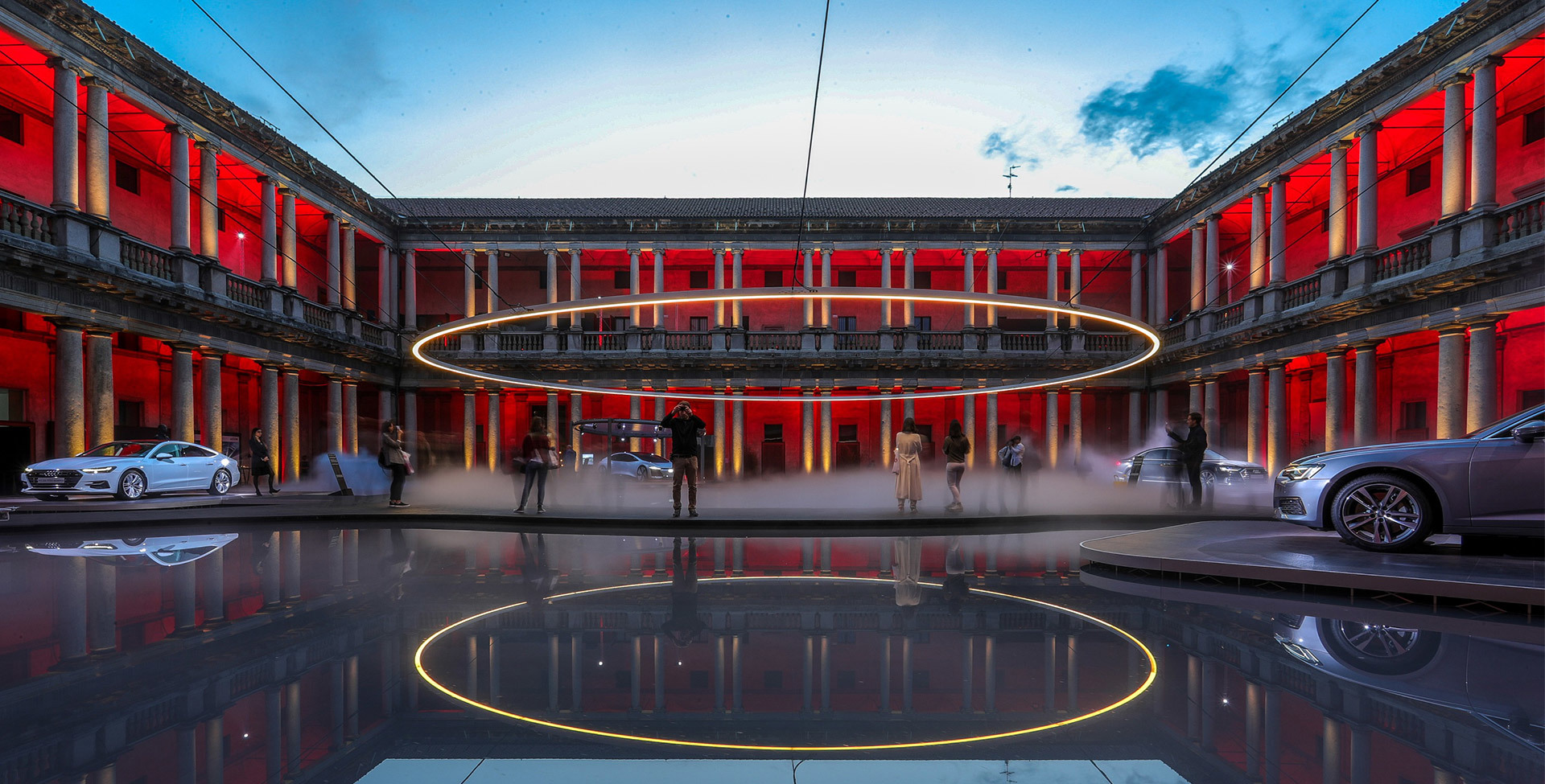Grande installazione ad anello luminoso sospesa in un cortile storico, immerso in luce rossa, con auto e visitatori riflessi sul pavimento bagnato.