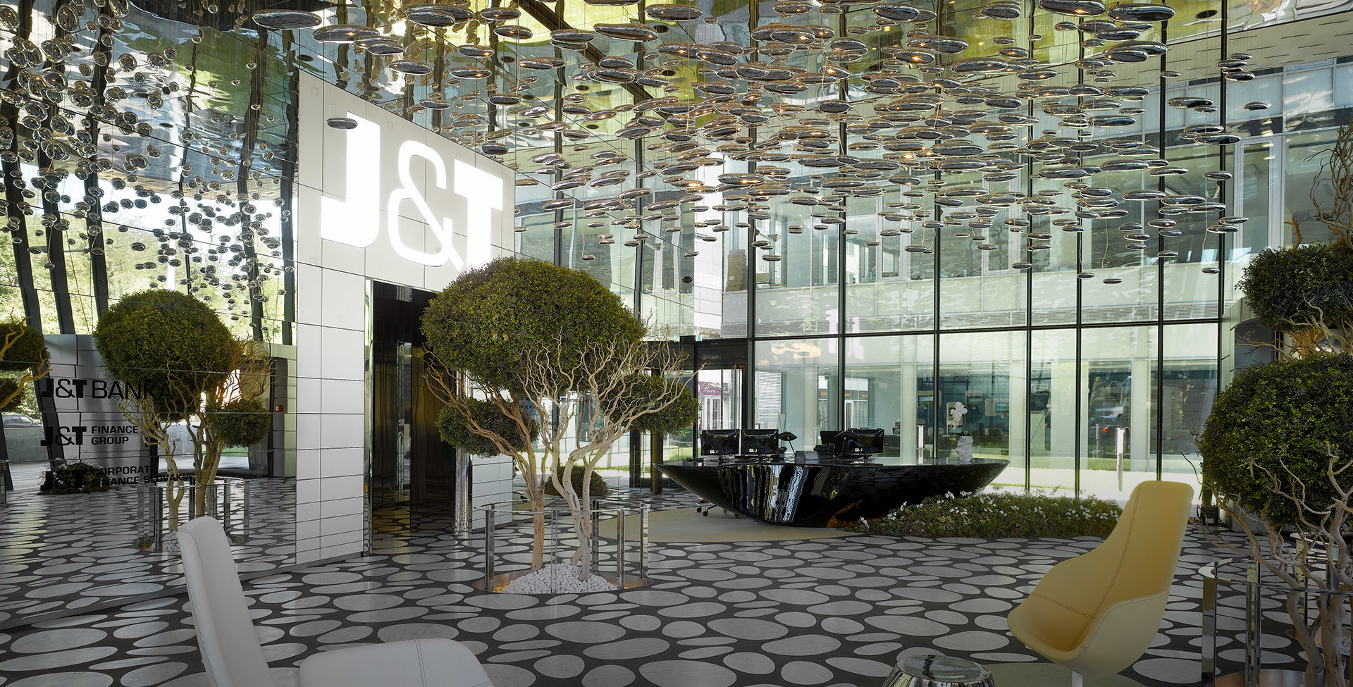 Lobby della banca J&T con lampade a sospensione Artemide che formano un'installazione a soffitto riflettente sopra alberi curati e arredi moderni