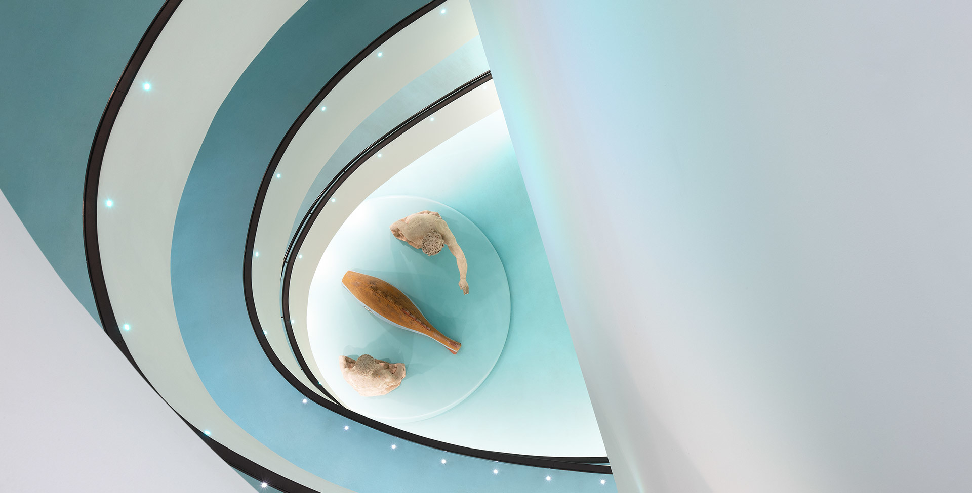 Aerial view of a spiral staircase with teal and white tones, small ceiling spotlights, and three sculptural objects at its center