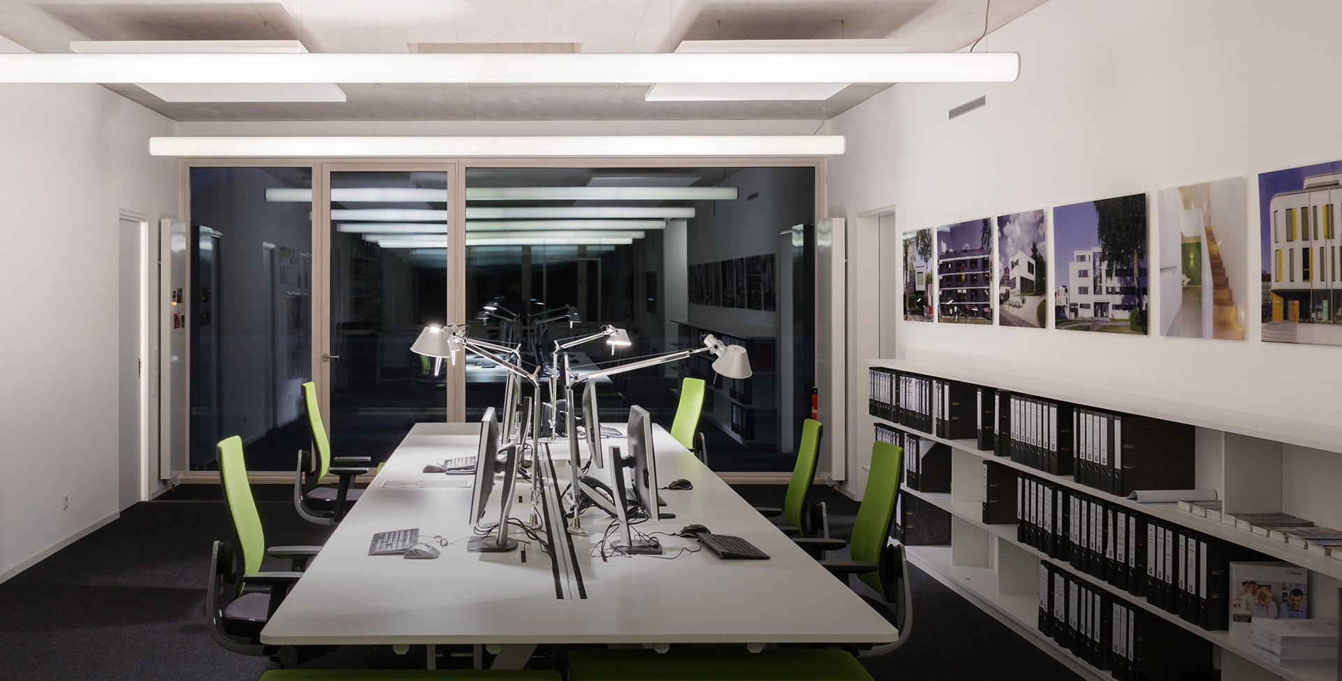 Modern office workspace with white desks, green chairs, Artemide Tolomeo desk lamps, and recessed linear ceiling lights illuminating the room.