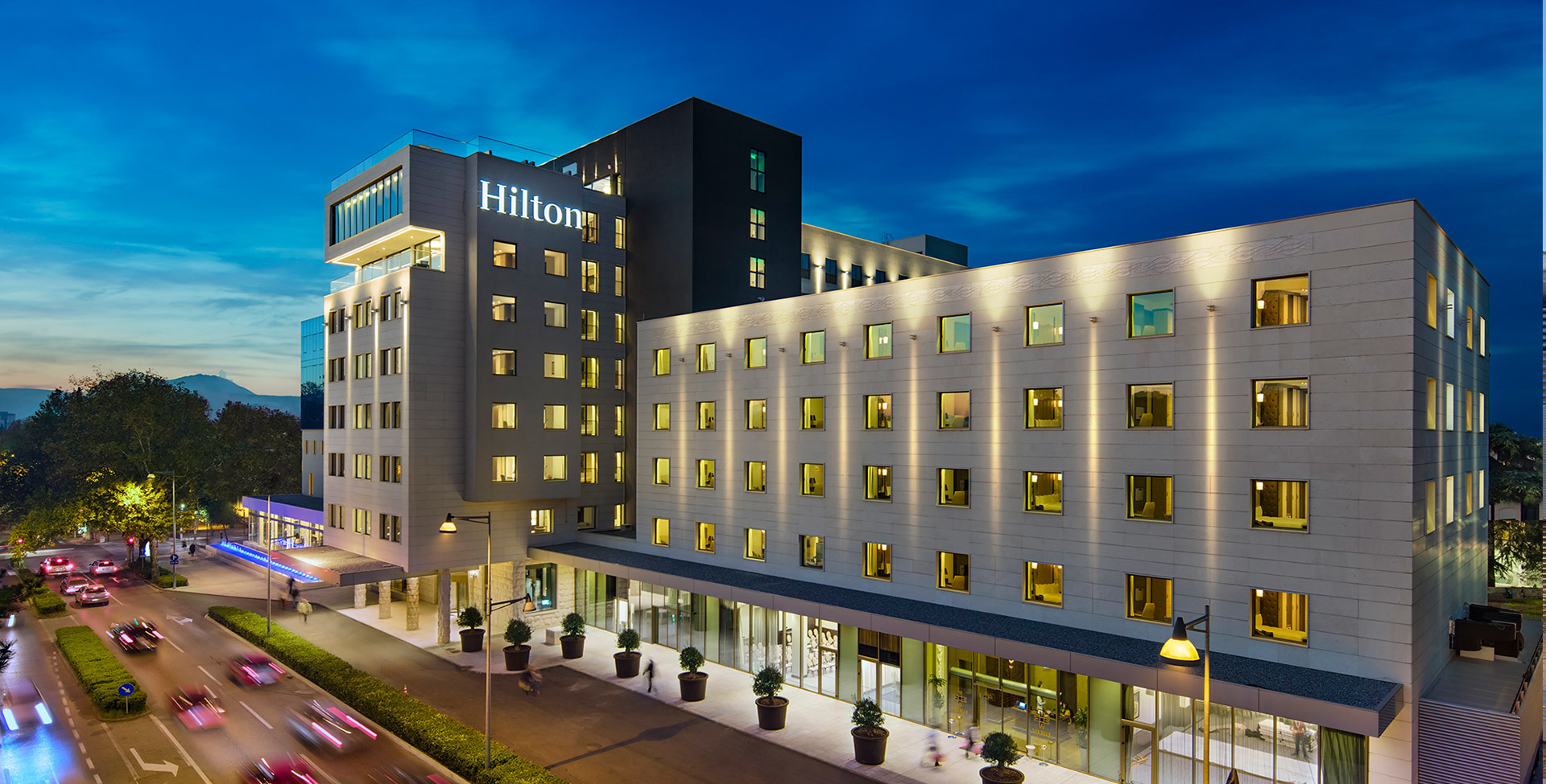 Hilton hotel exterior at dusk, illuminated facade with warm golden lighting highlighting the modern multi-story building.