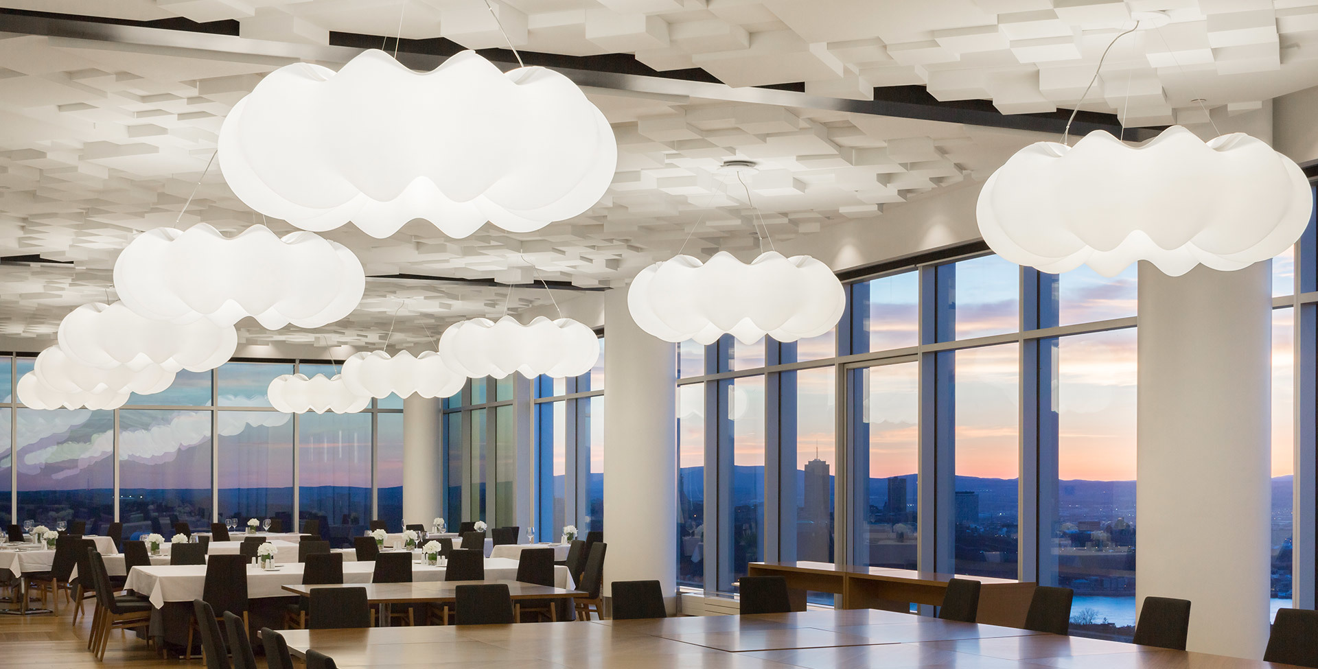 Restaurant with multiple Artemide cloud-shaped white pendant lights suspended from ceiling, floor-to-ceiling windows with city sunset view