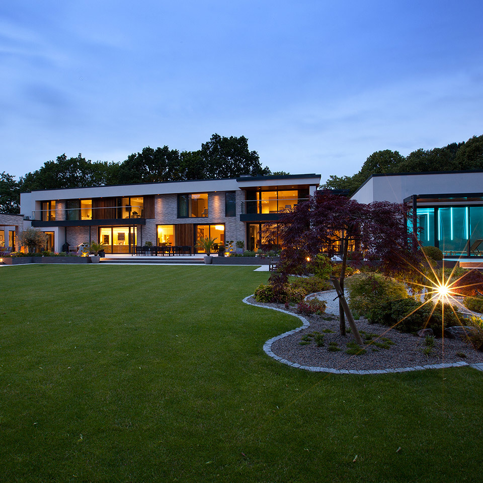 Modern villa at dusk with warm interior lighting and garden spotlights illuminating landscaped lawn and Japanese maple trees