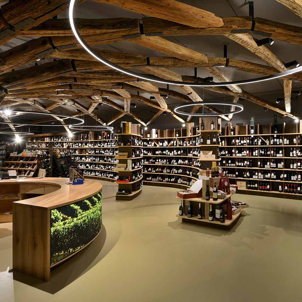 Interno di enoteca con lampade circolari a sospensione Artemide tra travi rustiche in legno, che illuminano scaffali curvi con bottiglie.