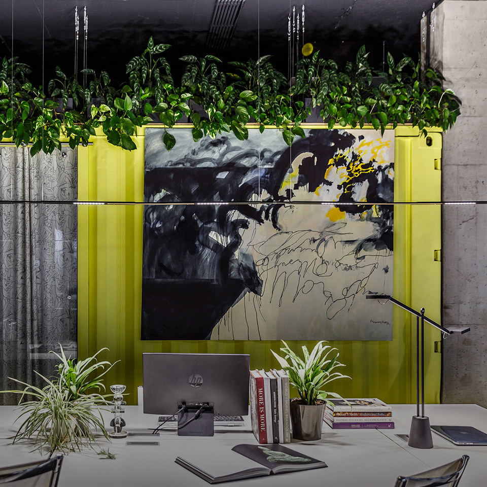 Modern workspace with Artemide desk lamp, laptop, books, plants, abstract artwork on yellow-framed wall, and hanging greenery above.