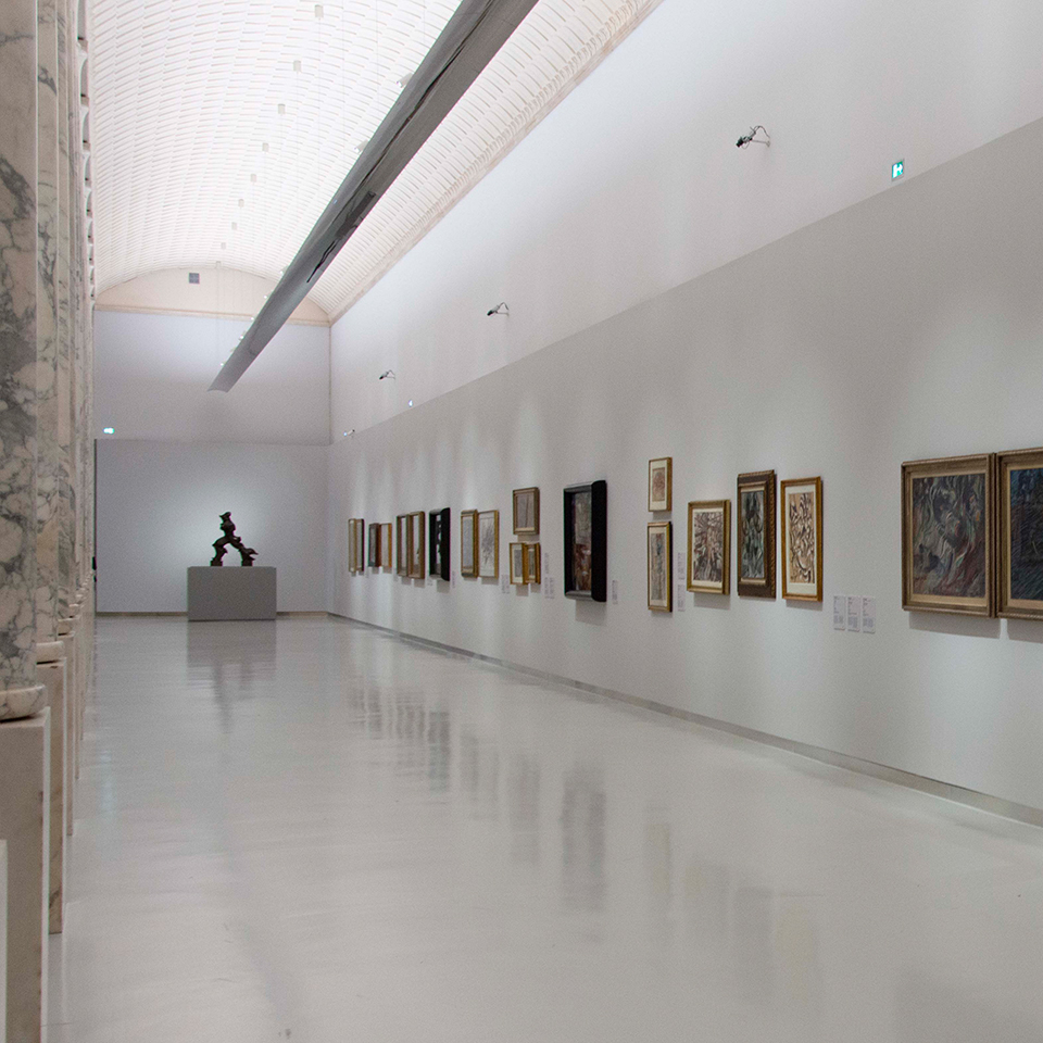 Museum gallery with white walls displaying framed artworks, lit by directional spotlights on ceiling tracks, polished reflective floor.