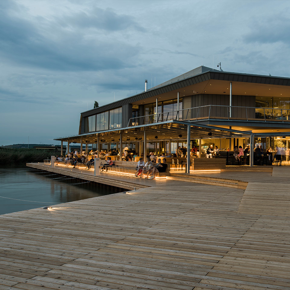 Waterfront restaurant at dusk with warm Artemide lighting illuminating a wooden deck terrace over calm water, guests dining outdoors