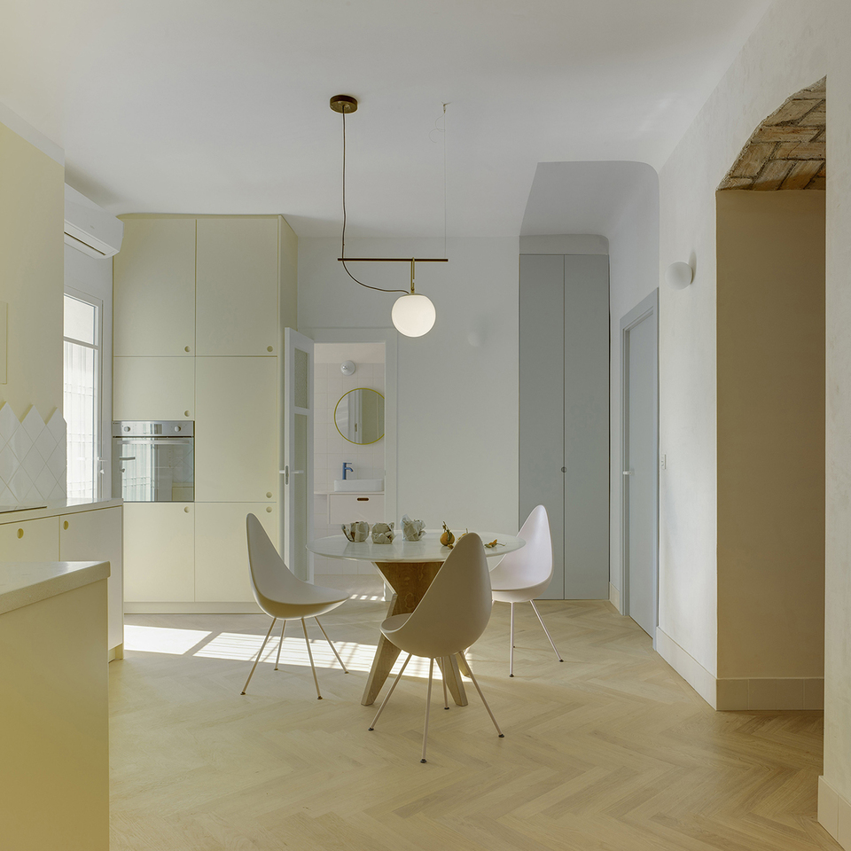 Vista della cucina con zona pranzo, pavimento in legno a spina di pesce e arredi in tonalità crema, il tutto illuminato da una lampada a sospensione.